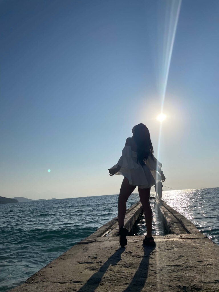Viajera disfrutando de una vista panorámica del mar Egeo desde un puerto, con el sol iluminando su rostro. Un pescador aparece en el fondo, mientras la mujer observa hacia arriba, absorbiendo la tranquilidad del paisaje mediterráneo.